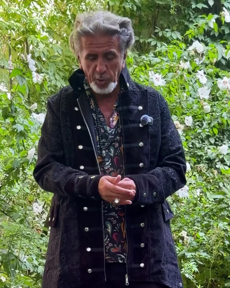 Hamid trägt den schwarzen Mantel Marven offen über einem bunten Hemd, im Stoff ein dunkles Paisleymuster